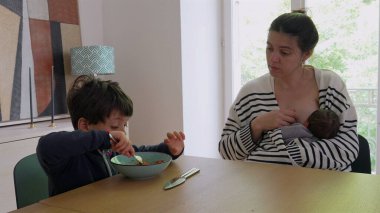 Anne, yeni doğan bebeği emzirirken ve daha büyük çocuğu yemek masasında beslerken, ebeveynlik ve günlük işleri bir evde dengelemenin zorluklarını göstermektedir.