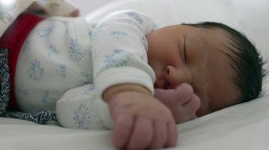 Yeni doğmuş bir bebek, bir eli hafifçe yüzüne yakın kıvrılmış bir şekilde bir yatakta rahat bir şekilde uyuyor. Yavruların sakin ifadesi ve rahat duruşu huzurlu ve huzurlu bir anın altını çiziyor.