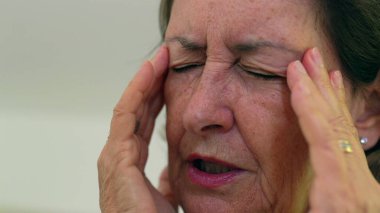 Kıdemli kadın gözlerini sıkıca kapatıyor ve iki eliyle tapınaklara masaj yapıyor. Acıyı ya da stresi azaltmaya odaklanıyor. Kırışıklıklar ve ifade, duygusal yükü vurgular