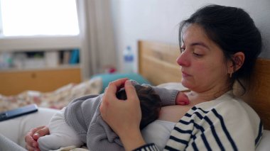 Anne, uyuyan bebeği göğsünde yatağında dinlenirken, gözleri bir an için sessiz bir yansımayla kapanıyor. Anne ve bebek arasında paylaşılan huzur ve huzur.