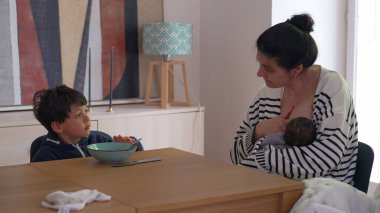 Anne yeni doğan bebeği emzirirken, büyük oğlu yemek masasında oturuyor, elleri kasesinin üzerinde, düşünceler içinde kaybolmuş, sessiz, düşünceli bir aile anını sergiliyor.