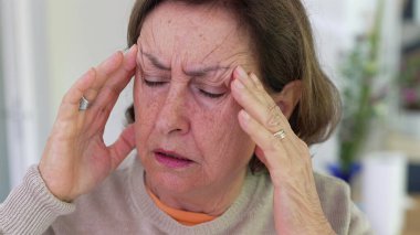 Büyük stres altındaki yaşlı kadın, parmaklarıyla tapınaklara masaj yapıyor, gözleri odaklı, yoğun endişe ve zihinsel yük altında. Detaylı ifade, duygusal gerginliğin etkisi veya fiziksel acı