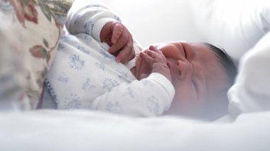 Yeni doğmuş bebek yatakta yatıyor, elleriyle jestler yapıyor ve yüzünü buruşturuyor. Bebek yumuşak yastıklarla çevrili, yatak odasında rahat ve huzurlu bir atmosfer yaratıyor..