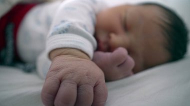 Yeni doğan bebek bir eli açık, diğer eli yüzü yakın bir şekilde bir yatakta huzur içinde uyuyor. Çocuğun sakin ifadesi ve rahat duruşu bir huzur ve güvenlik anını vurguluyor.