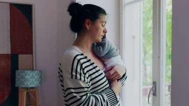 Anne pencerenin kenarında durmuş, yeni doğmuş bir bebek omuzunda, dışarıya bakıyor. Sahne, anne ve bebek arasındaki sakinlik, bağ ve huzur dolu anları aktarır.
