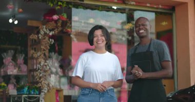 Çiçekçinin önünde yan yana duran iki dükkan sahibi kameraya gülümsüyor. Renkli vitrinler ve çiçek vitrinleri arkasında.