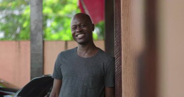 Afrika kökenli bir adam duvara yaslanırken sıcak bir şekilde gülümsüyor, kentsel arkaplan ve yeşillik yakınlarında günlük kıyafetlerle neşeli bir ifade sergiliyor.