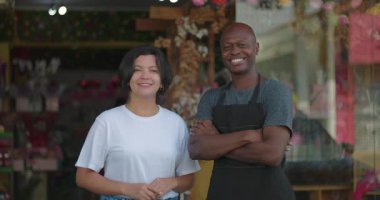 Çiçekçi dükkanının önünde duran küçük işletme sahipleri, beyaz gömlekli, arka planda mağaza önü açık, kolları bağlı, gülümseyen bir adam.