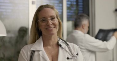 Steteskoplu gülümseyen kadın doktor profesyonel tıp bürosundaki kameraya bakarken, erkek meslektaşı arka plandaki beyin taramasını inceliyor.