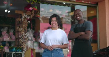 Çiçekçinin önünde gururla gülümseyen iki iş arkadaşı, kolları rahat, kameraya bakıyor, çiçeklere bakıyor ve arka planda canlı mağazalar var.