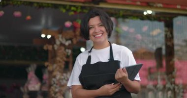 Çiçekçi çiçekçinin önünde elinde tablet tutarken ışıl ışıl gülümsüyor, çiçekçi ortamında pozitif enerji yayıyor ve iş yapmaya hazır.