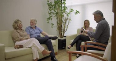 Klinikteki bekleme salonundaki bir grup hasta, koltuklarda oturuyor ve yaşlı çiftler de dahil olmak üzere cep telefonları kullanıyorlar.