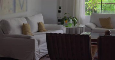 Beyaz kanepeli parlak oturma odası, kahve masası, kapalı salonda bitkiler ve yeşil manzaralı geniş pencere, doğal ışık ve sıcak tonlu rahat modern alan.