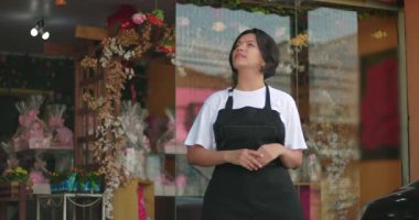 Çiçekçi dükkanının önünde kendine güvenen, kollarını kavuşturmuş, kararlılıkla, çiçeklerle ve mağaza vitrinleriyle yukarıya bakan bir kadın.