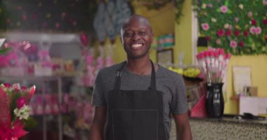 Tezgahın arkasındaki çiçekçide gülümseyen erkek çiçekçi. Etrafı buketlerle, çiçeklerle ve neşe dolu iş ortamlarıyla çevrili.