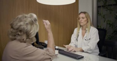Modern klinik ofisinde, masaüstü bilgisayarı ve sakin, profesyonel ayarlarıyla tıbbi danışma sırasında yaşlı kadını dikkatle dinleyen kadın doktor.