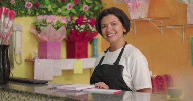 Çiçekçide gülümseyen kadın defterleri açık, sarılı çiçeklerle ve renkli atölyelerde gül buketleriyle çevrili.