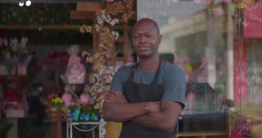 Dükkanın önünde kolları çapraz duran çiçekçi, tarafsız ifade, çiçeklerin ve vitrinin canlı arkaplanı, küçük işletme temsilcisi