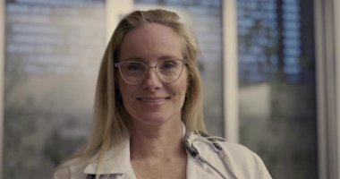 Steteskoplu neşeli kadın doktorları kapatın. Ofiste kameraya gülümsüyor, cam kaplama arka planda.