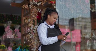 Çiçekçi gülümsüyor ve çiçekçi dükkanının dışındaki telefona bakıyor. Çiçek aranjmanları ve dekoratif butik girişi yakınlarındaki mesajları kontrol ederken tatilin tadını çıkarıyor.