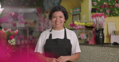 Çiçekçinin arkasındaki kameraya gülümseyen çiçekçi renkli düzenlemeler, dekoratif buketler ve perakende satış alanında hediye eşyalar ile çevrili.