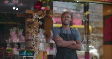 Çiçekçinin önünde gururla duran çiçekçi, kollarını kavuşturmuş, tarafsız ifade, renkli çiçek aranjmanları ve arka planda hediyeler.