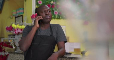 Afro-Amerikalı girişimci çiçekçi dükkanında telefonla konuşuyor, kendine güvenen bir duruş ve odaklanmış bir ifadeyle işi tartışıyor.