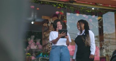 Çiçek butiğinin önünde elinde tablet tutarken gülümseyen ve sohbet eden kadınlar, iş ortamında rahat takım çalışmasının keyfini çıkarıyorlar.