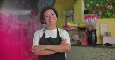 Çapraz kollu bir çiçekçi ve çiçekçi dükkanında sıcak bir gülümsemeyle durmakta. Renkli buketler, çiçek aranjmanları ve canlı dekoratif eşyalarla dolu.