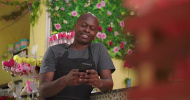 Tezgahın arkasında telefon kullanırken gülümseyen Afrikalı Amerikalı çiçekçi renkli çiçekçide neşeli iş anlarının tadını çıkarıyor.