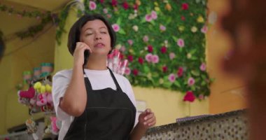 Çiçekçi telefonda konuşuyor ve renkli çiçekçide tezgahın arkasında notlar alıyor. Arka planda yapay güllerden ve hediye malzemelerinden oluşan dekoratif bir duvar var.