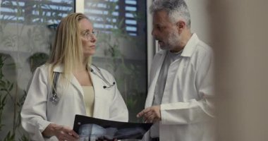 Kadın doktor önplanda güvenle dururken, erkek meslektaşı tıbbi danışma sırasında arka plandaki röntgeni inceliyor.