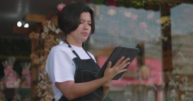 Çiçekçinin önündeki tableti kullanan çiçekçi, çiçekçi dükkanının önünde dikilirken dijital görevlere ve planlamaya odaklanmış.