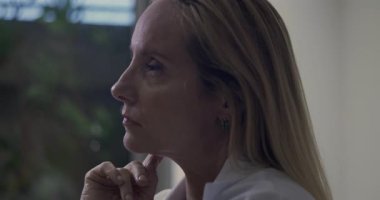 Yan profildeki kadın doktor, elleri çenesinin altında, gözleri klinik ortamda derin bir yansıma anında ileri bakıyor.