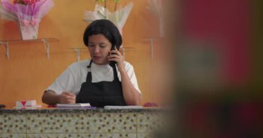 Çiçekçi, çiçekçi tezgahında gülümseyerek notlar alıyor. Çiçek hediyeleri, gül aranjmanları ve paketlenmiş çiçek kutularıyla dolu.