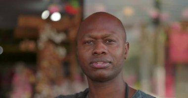 Tarafsız bir ifadeyle çiçekçinin yakın plan portresi, bulanık çiçekçi arka planının önünde duruyor, içsel ve sakin bir an.