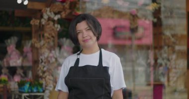 Çiçekçinin önünde yüzünde yumuşak bir ifadeyle duran, süs çiçekleri ve hediye sepetleriyle çevrili, barışçıl ve misafirperver küçük iş atmosferini yansıtan.