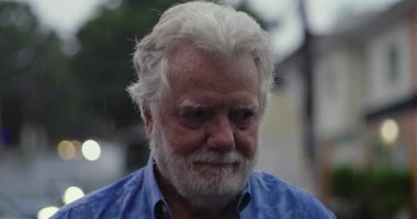 Yağmurda acı tatlı gülümseyen yaşlı bir adam, gözyaşı dolu gözler ve hüznü ve kabulü yansıtan şefkatli bir ifade.