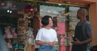 Çiçekçinin önünde konuşan iki dükkan sahibi, gülümseyen ve arkadaşça sohbet eden, çiçeklerle dolu canlı bir mağaza.
