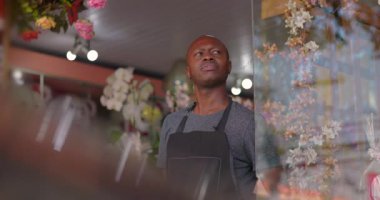 Afro-Amerikalı bir adam çiçekçi dükkanının penceresinden dışarı bakıyor. Etrafı çiçeklerle ve hediye eşyalarıyla çevrili.
