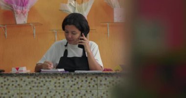 Çiçekçinin arkasında çiçek kutuları, çiçek hediyeleri ve arka planda gül süslemeleriyle notlar alan ve telefonda konuşan kadın.