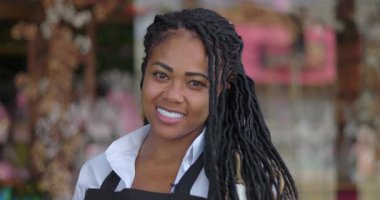 Uzun örgülü saçlarıyla gülümseyen çiçekçiye yaklaş ve mevsimlik süslemelerle dolu çiçekçinin önünde kameraya sıcaklıkla ve gururla bak.