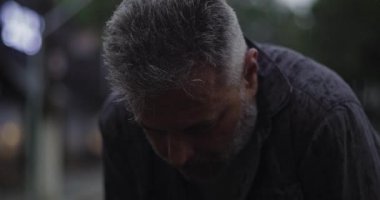 Orta yaşlı bir adam, başı yağmurda öne eğik, derin bir üzüntü ya da yorgunluk ifade ediyor.