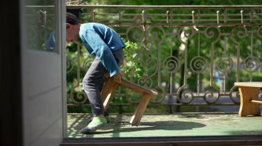 Genç bir çocuk balkonda küçük bir taburede oturuyor, bacaklarını açmış, dışarıda huzurlu bir anın tadını çıkarıyor. Güneşin tadını çıkaran bir çocuğun rahat ve kaygısız havası.