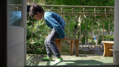 Genç bir çocuk balkonda küçük bir taburede oturuyor, bacaklarını açmış, dışarıda huzurlu bir anın tadını çıkarıyor. Güneşin tadını çıkaran bir çocuğun rahat ve kaygısız havası.