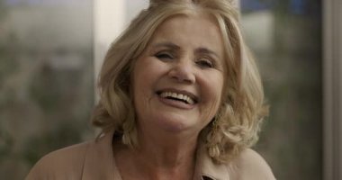 Sarışın ve anlamlı gözleri olan gülümseyen yaşlı bir kadın doğrudan kameraya bakıyor. Ev ortamında sıcaklığı, mutluluğu ve kendine güveni aktarıyor.