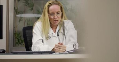 Kadın doktor masasında oturuyor. Yorgunluk ve duygusal yorgunluk gösteriyor. Gözler kapalı ve elleri klinikte iş sırasında yüzün üzerinde.