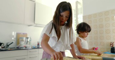 Mutfakta kardeşler yemek hazırlıyor, kız ve oğlan yemek pişirmeye odaklanıyor, aşçılık faaliyetleri, aile ilişkileri, arka planda anne, ışık dolu mutfak.