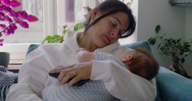 Anne bebeğini kucağında, bebek yüzüne dokunurken yavaşça kanepede dinleniyor. Huzurlu bir ev ortamında samimi bir yakınlık ve bağ anı.