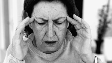 Şiddetli baş ağrısı ile mücadele eden yaşlı bir kadın, gözle görülür konsantrasyonda şakaklara masaj yapıyor. Yüz hatları ve yüz ifadeleri yoğun acıyı, zihinsel gerginliği, duygusal sıkıntıyı yansıtır.